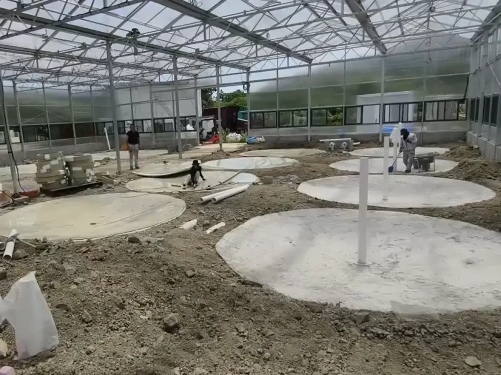 Workers preparing circular concrete pads inside a greenhouse-style building.