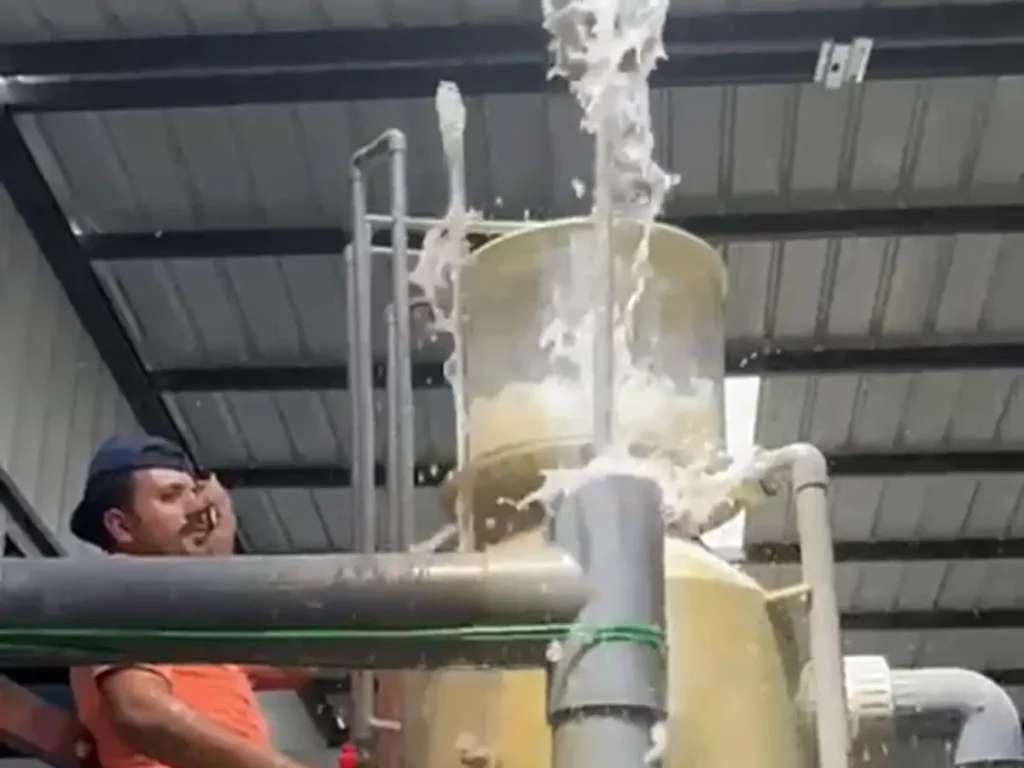 A person beside a vertical vessel with visible water flow and connected pipes.